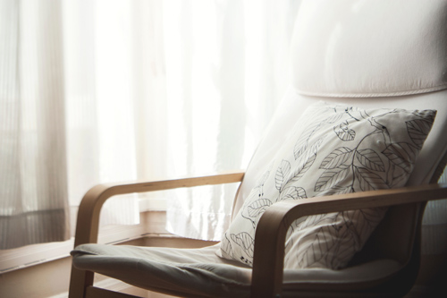 Picture of a wooden chair with fitted cushions and a single extra cushion with leaf outline pattern, in a soft lit room with pale curtains in background. Taken by Kari Shea on Unsplash
